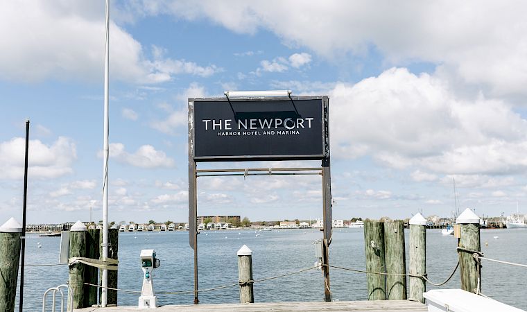 A marina pier sign reads “The Newport Harbor Hotel and Marina,” with blue sky and water in the background.