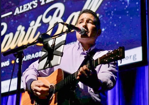 A man sings and plays an acoustic guitar at a stage during a live &ldquo;Alex&rsquo;s Team&rdquo; event, with a starry banner behind him.