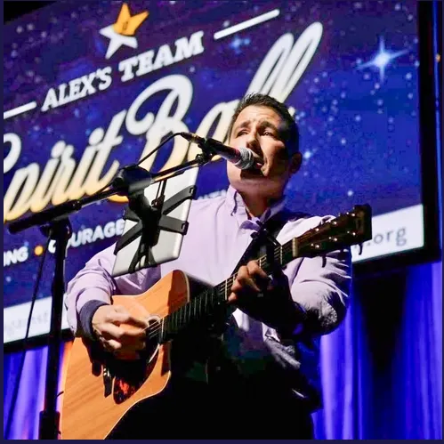 A man sings and plays an acoustic guitar at a stage during a live &ldquo;Alex&rsquo;s Team&rdquo; event, with a starry banner behind him.