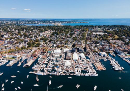An aerial view of a coastal town and busy harbor with many sailboats and yachts docked along the marina, overlooking the sea.