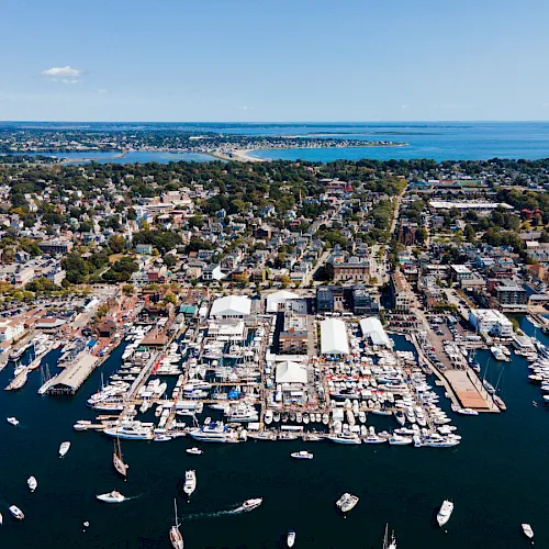 An aerial view of a coastal town and busy harbor with many sailboats and yachts docked along the marina, overlooking the sea.