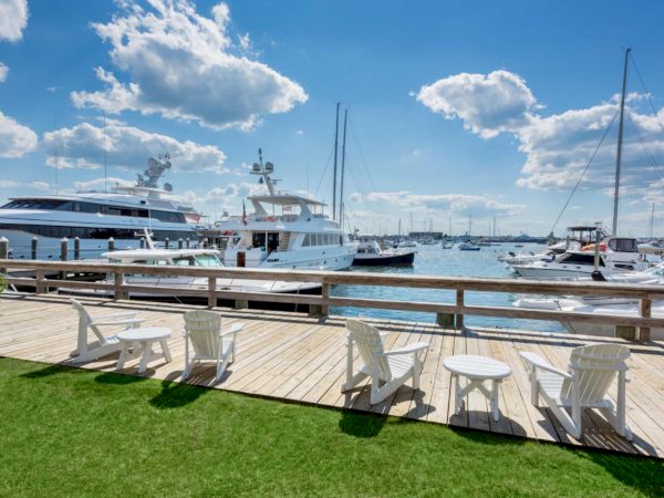 A sunny marina with docked yachts and masts, plus white patio chairs on a wooden pier.