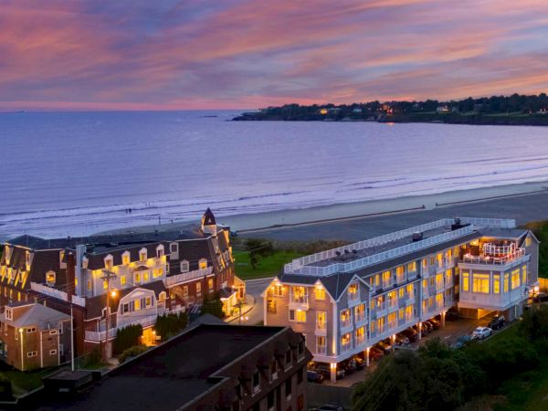 A seaside hotel overlooks a calm beach at dusk, with warm lights and a pink-purple sky stretching over the water.