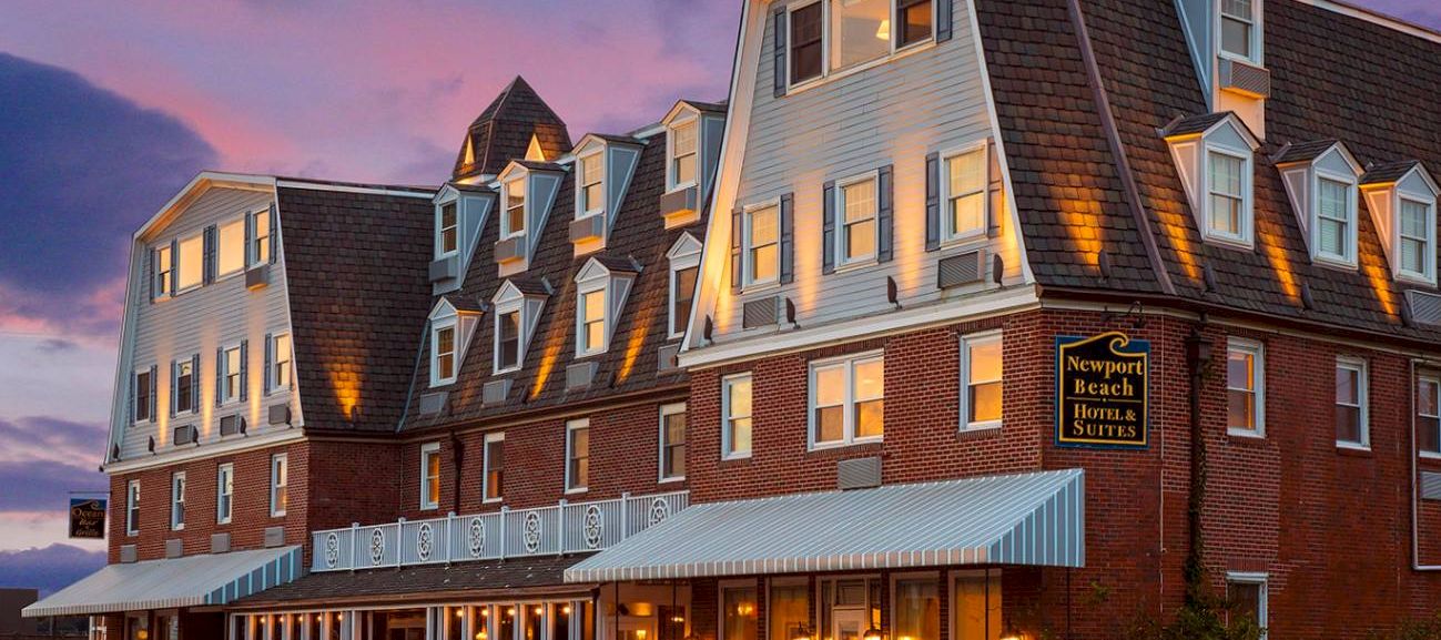 A brick hotel building with dormers and lit windows at dusk, featuring a &ldquo;Newport Beach Hotel & Suites&rdquo; sign out front.