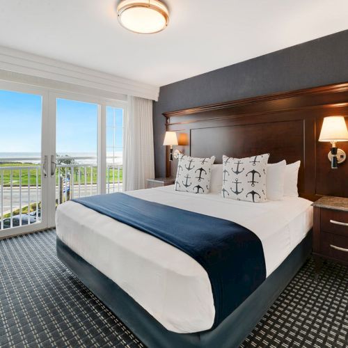 A seaside hotel bedroom with a large bed, navy-and-white bedding, bedside lamps, and ocean-view balcony doors.
