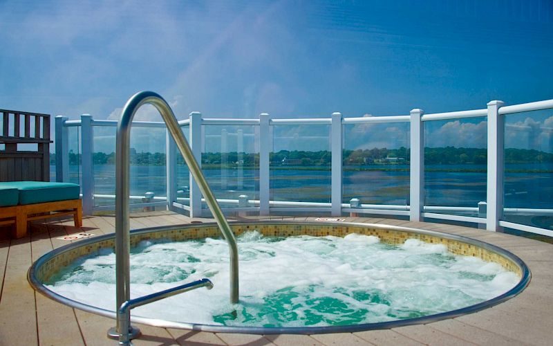 An outdoor hot tub with bubbling water sits on a deck behind glass railing, overlooking a calm lake under a blue sky.