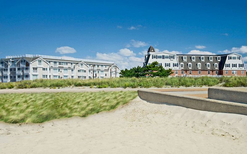 A seaside resort of long, multi-story buildings with windows sits on a sandy beach under a blue sky.