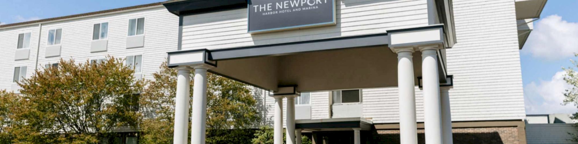 White hotel building with columns and a sign reading &ldquo;THE NEWPORT&rdquo; for Harbor Hotel and Marina under a clear blue sky.