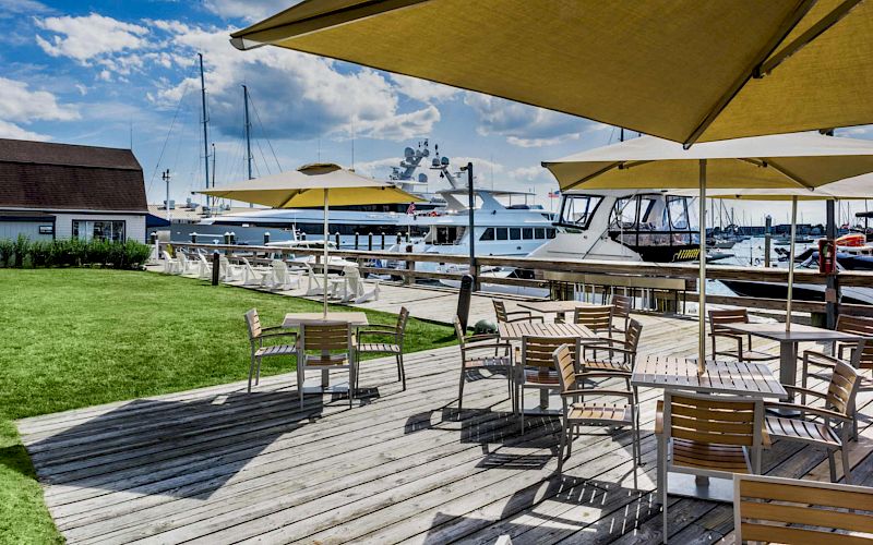 Outdoor marina deck with chairs under yellow umbrellas, overlooking moored yachts and a large ship.