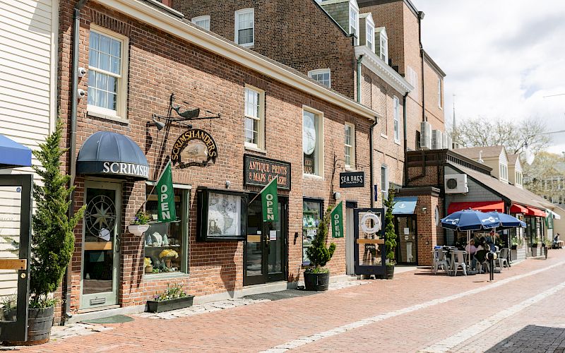 A brick shop-lined street with signs for Scrimshanders, antique prints, and sea bags, plus open banners and outdoor tables.