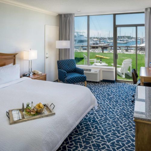 A hotel room with a large bed and seating, with big windows overlooking a marina of sailboats and chairs.
