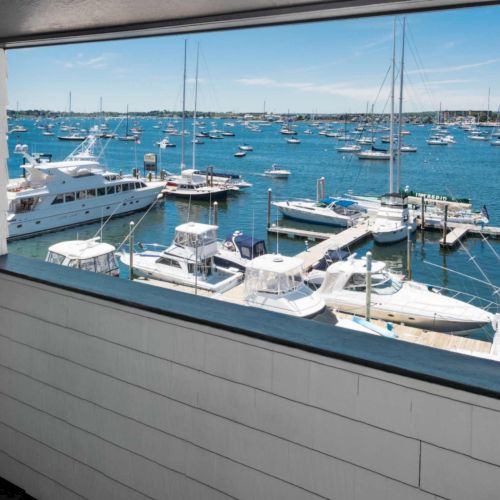 A sunny marina view with many sailboats and yachts docked along a pier, seen from a building balcony.