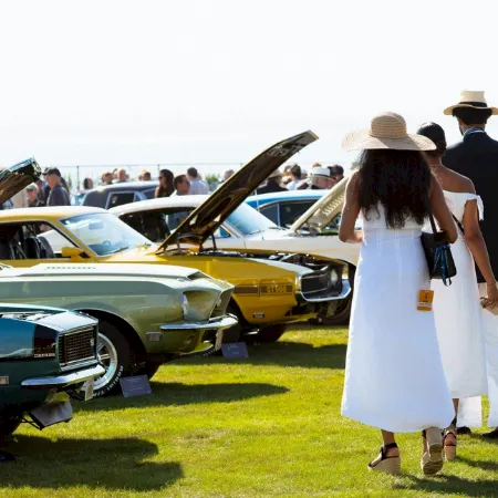 People walk through a sunny car show, with classic cars on display and hoods open in rows.