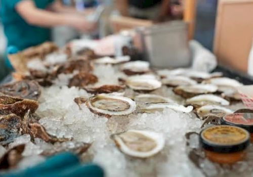 A vendor&rsquo;s table of oysters on crushed ice with lemon slices and sauce cups in a busy market.