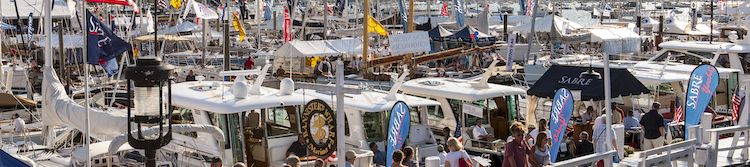 A busy marina packed with sailboats, flags, and people walking along the dock, with water and sail masts stretching into the distance.
