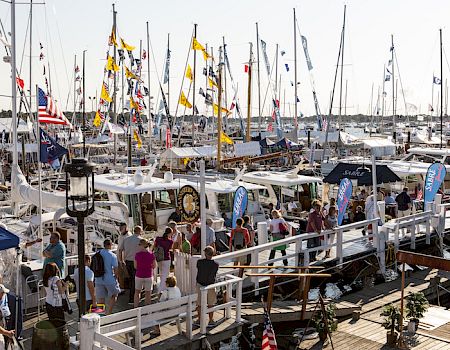 A busy marina packed with sailboats, flags, and people walking along the dock, with water and sail masts stretching into the distance.