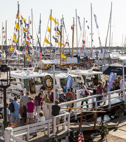 A busy marina packed with sailboats, flags, and people walking along the dock, with water and sail masts stretching into the distance.