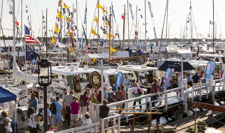 A busy marina packed with sailboats, flags, and people walking along the dock, with water and sail masts stretching into the distance.