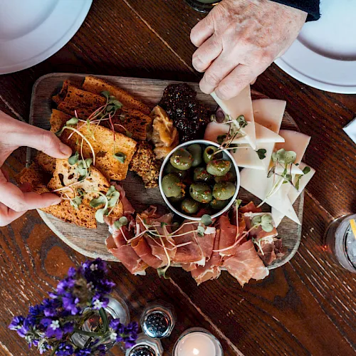 Hands arrange a charcuterie board with breads, cured meats, olives, sprouts, and drinks on a wooden table.